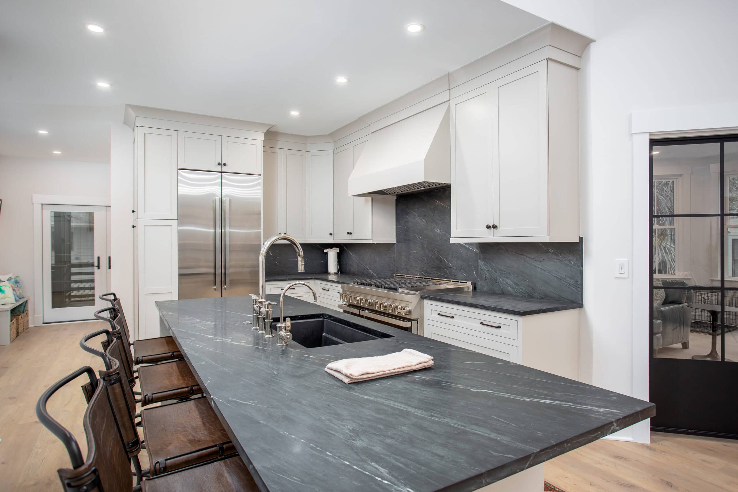 Isle Custom Homes Modern kitchen with white cabinets, stainless steel appliances, black stone countertops, an island with a sink, and bar stools.