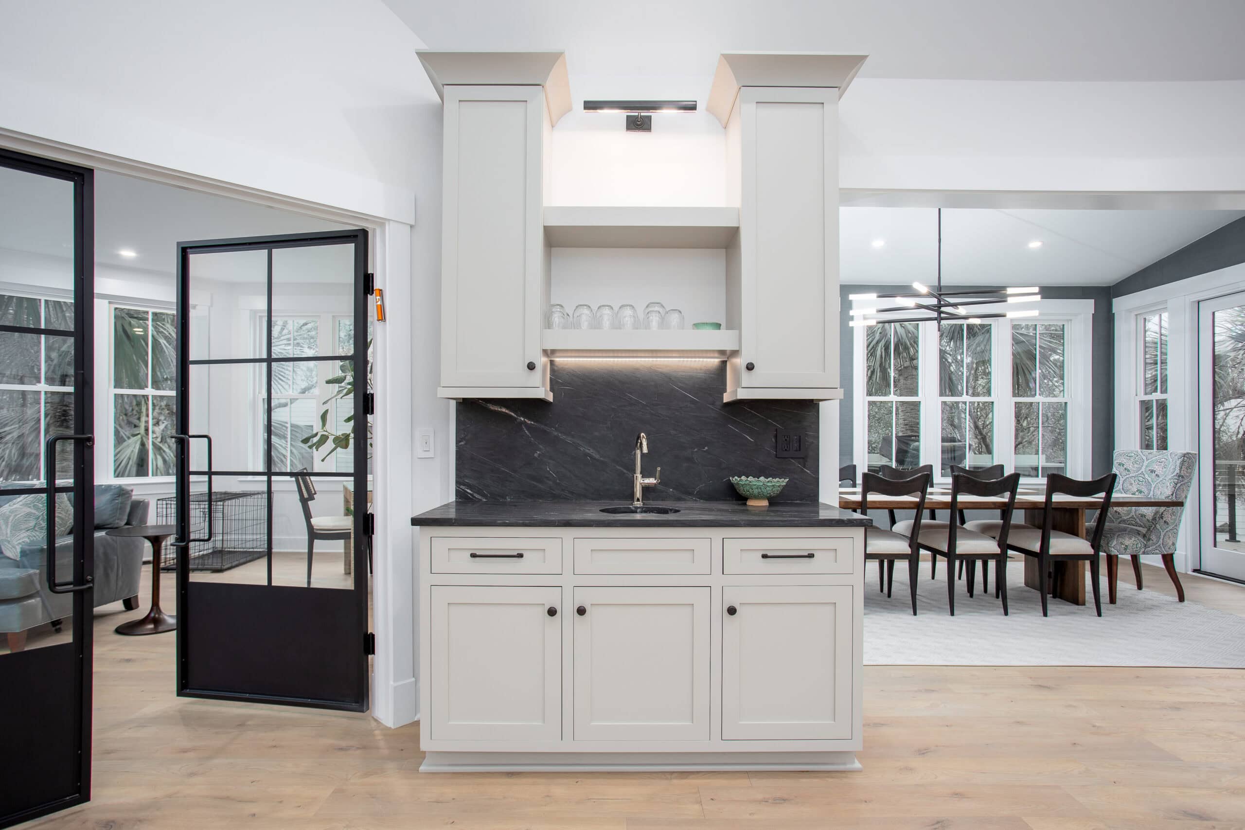Isle Custom Homes Modern kitchenette with white cabinets, black countertop and backsplash, open shelves with glassware, a sink, and a view into a dining room with a large table and chairs.