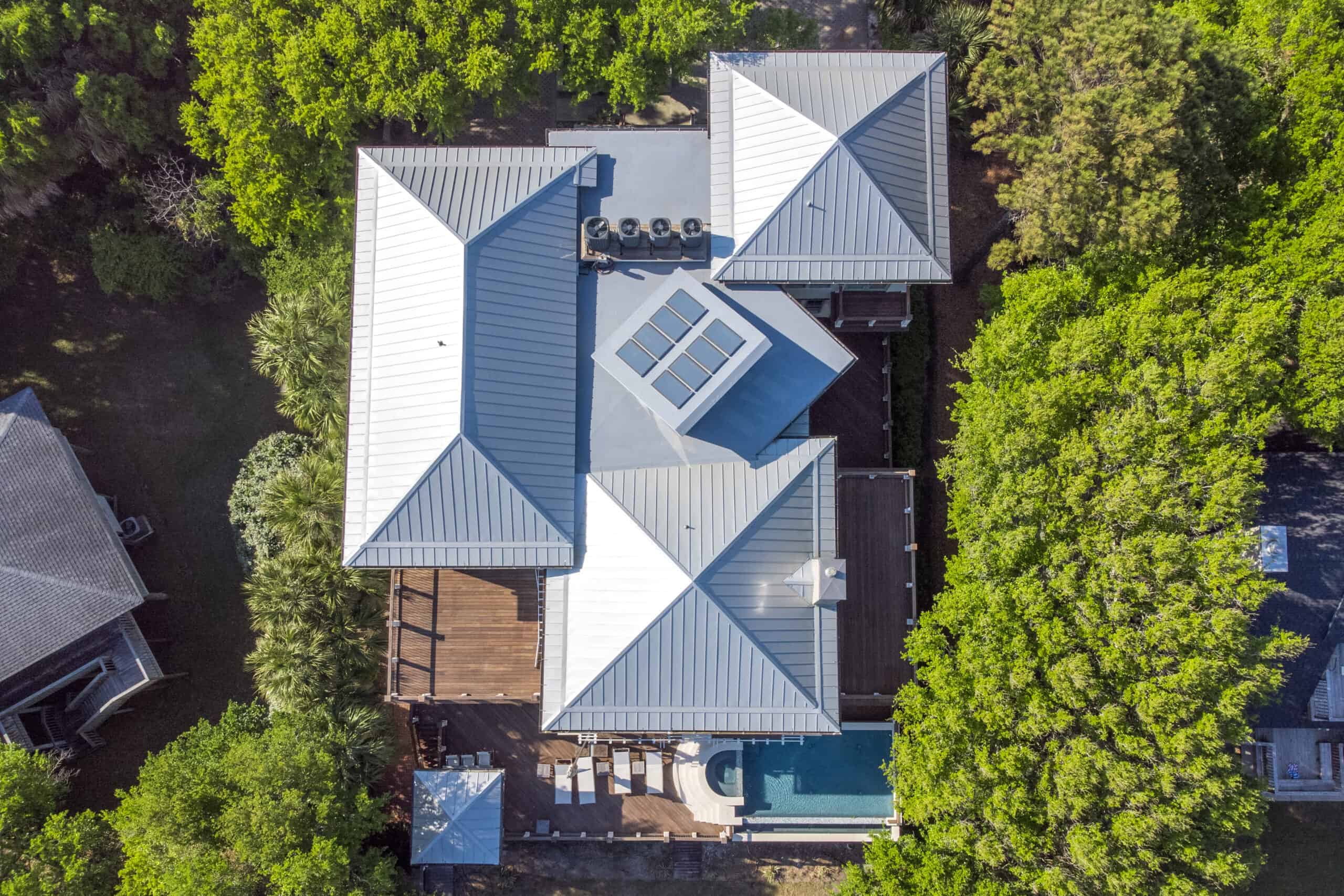 Isle Custom Homes Aerial view of a modern house with a gray metal roof, wooden deck, swimming pool, and surrounding green trees.