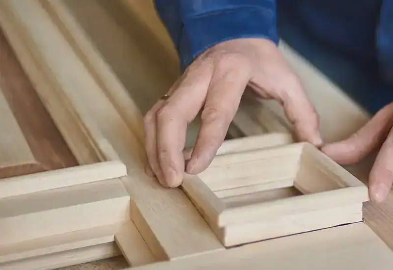 Isle Custom Homes A person assembling a wooden frame, placing a rectangular piece onto a larger wooden structure.