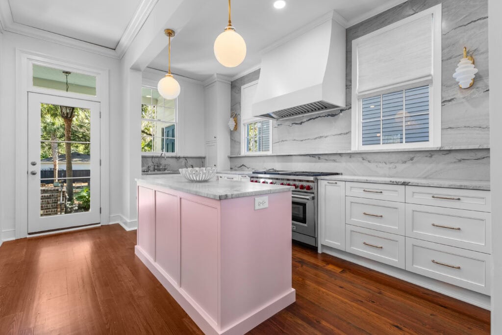 Isle Custom Homes Modern kitchen with white cabinets, a marble backsplash, a pink island, stainless steel appliances, and two globe pendant lights; wooden floor and large windows provide natural light.