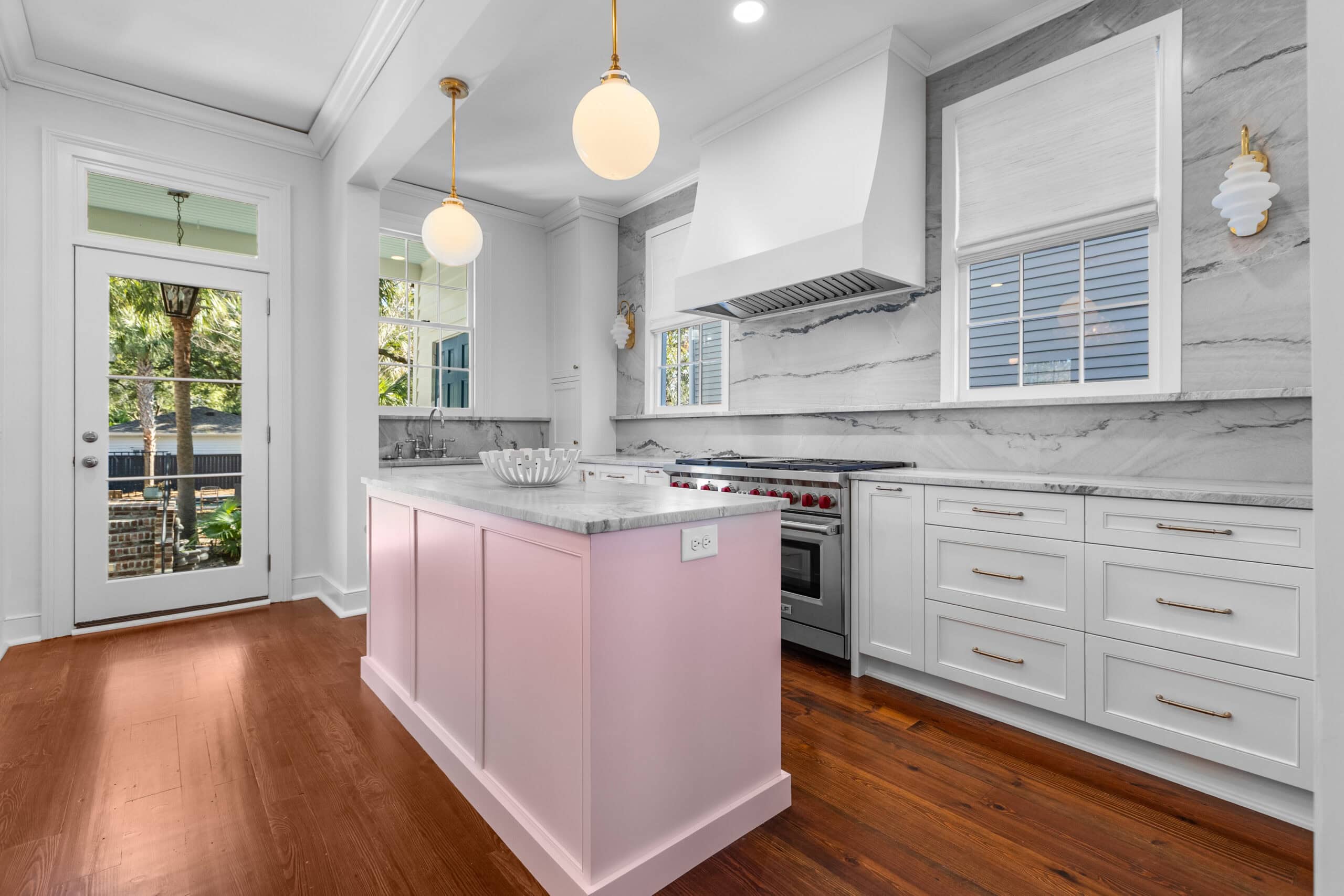 Isle Custom Homes Modern kitchen with white cabinets, a marble backsplash, a pink island, stainless steel appliances, and two globe pendant lights; wooden floor and large windows provide natural light.