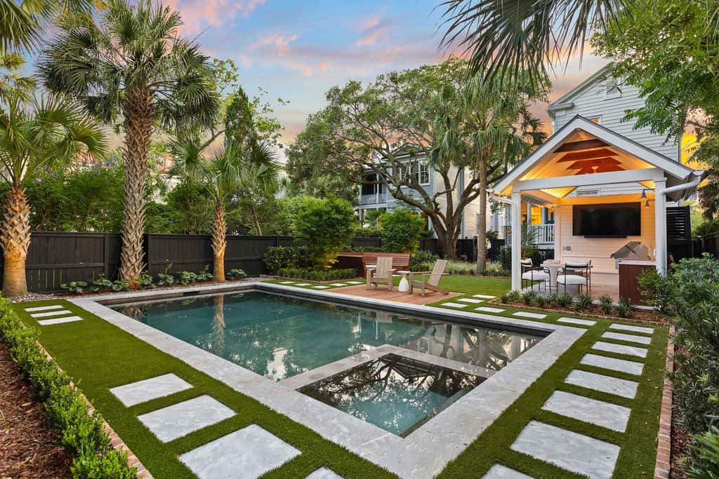 Isle Custom Homes Backyard with a rectangular swimming pool, surrounding stone pavers, lounge chairs, palm trees, and a covered outdoor seating area with a mounted TV.